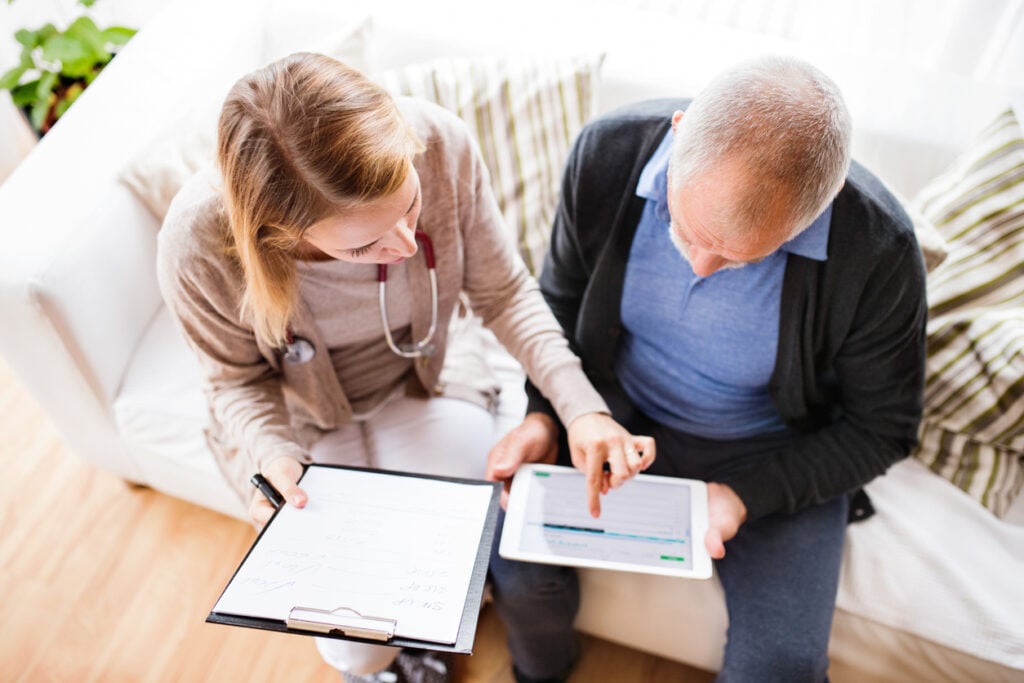 Healthcare worker visiting an elderly patient and helping him access his medical records online using a tablet device.