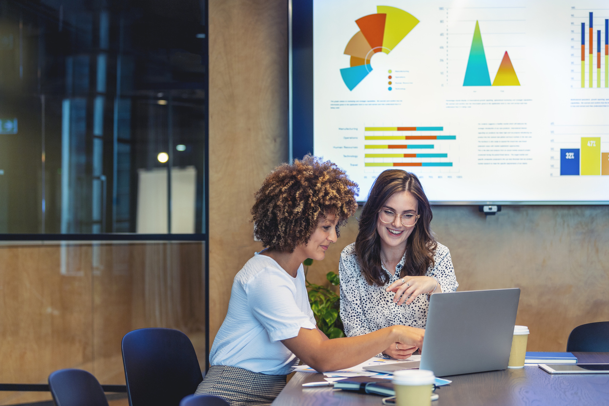 A photo of businesswomen colleagues working together on a laptop with a large screen on the background showing charts and graphs